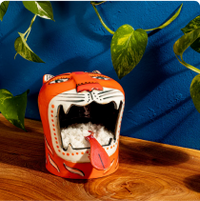 Orange tiger-face container with an open mouth showing spices inside, set on a wooden surface against a blue wall with trailing green vines.