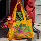 Person holding a bright yellow tote bag with colorful floral artwork and bold text, with a bouquet of flowers tucked beside it.